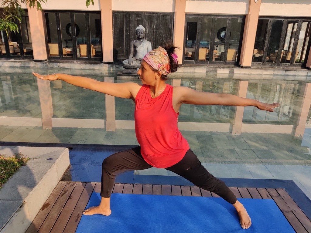 Sheetal Mody in Warrior II on a yoga mat beside a reflecting pool